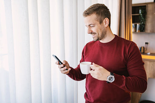 A man is standing at home and drinking coffee while smiling at the phone.