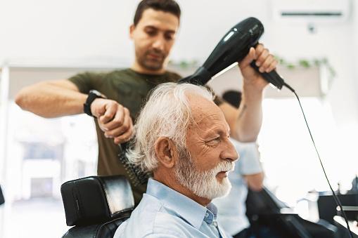 Senior man getting groomed at hairdresser with hair dryer while sitting in chair