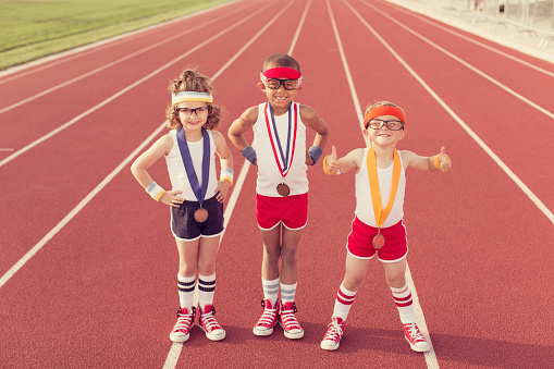 A group of three children dressed in vintage and retro workout attire are ready to show their running speed and agility on the track, albeit in a humorous way, winning medals along the way.