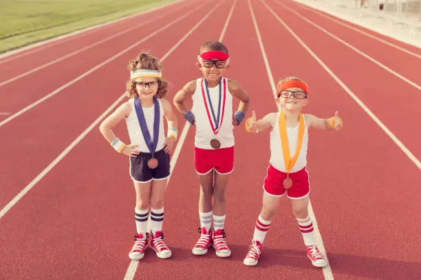 A group of three children dressed in vintage and retro workout attire are ready to show their running speed and agility on the track, albeit in a humorous way, winning medals along the way.
