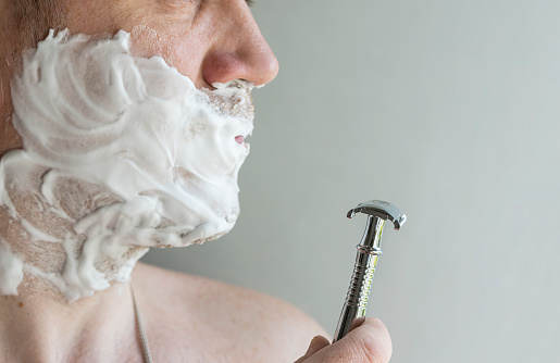 A handsome man shaves with a T-shaped metal chrome razor in natural light. copy space.
