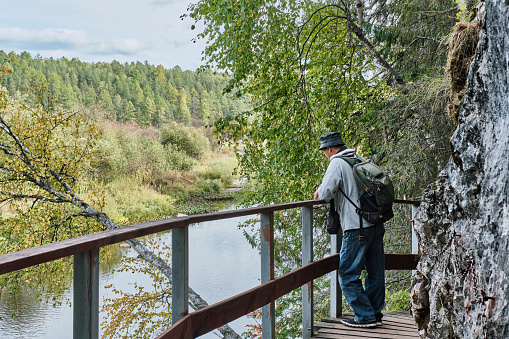 Back view of senior male hiker with backpack who is admiring summer nature landscape on wooden hiking trail near cliff. Nature park Deer streams, Sverdlovsk region, Russia. Serga River. Travel concept