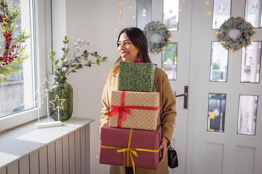 Young woman entering at home for Christmas. She is holding gift boxes and smiling to someone.