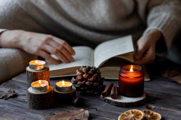 Cozy autumn or winter composition with aromatic candle, wool sweater, woman reading a book. Aromatherapy, home atmosphere of cosiness and relax. Wooden background close up.