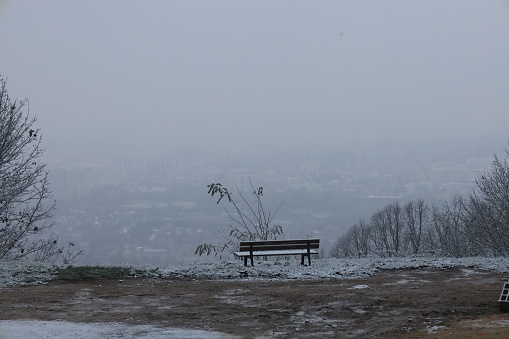 A wooden bench covered with snow on an empty rural road on a hill facing the foggy panorama of a city