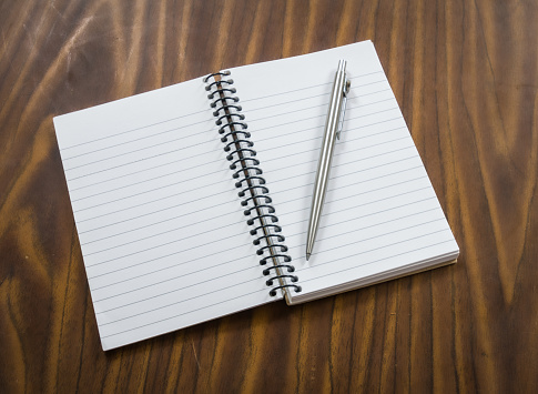 An open spiral bound notebook with a silver ball point pen lies open on a wood grained desk top.