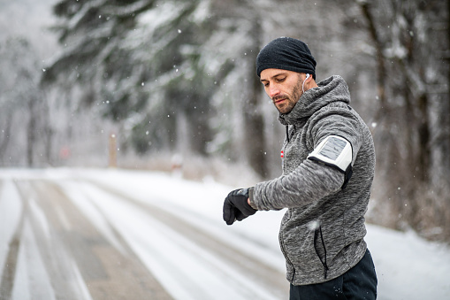 One man, an adult man exercises in the snowy forest in the fresh air, leads a healthy lifestyle, is dressed in sports clothes, looks at the watch that shows him all the training parameters