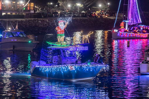Boats decorated with Christmas holiday lights makes its way around a harbor, with colorful lights reflected in the water.