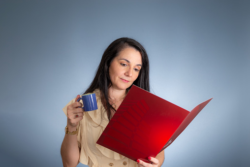 Portrait of female professional reading a document and drinking coffee. Isolated against light blue background.