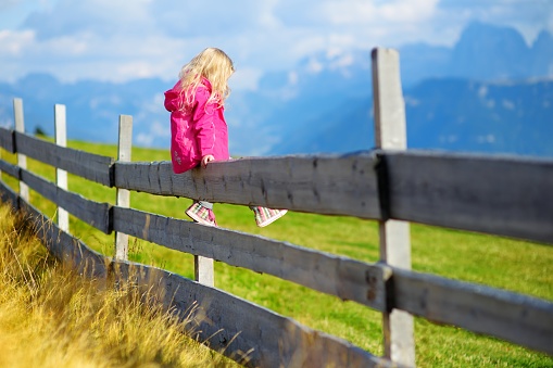 Cute little girl sitting on wooden fence admiring beautiful landscape in Dolomites mountain range, South Tyrol province of Italy