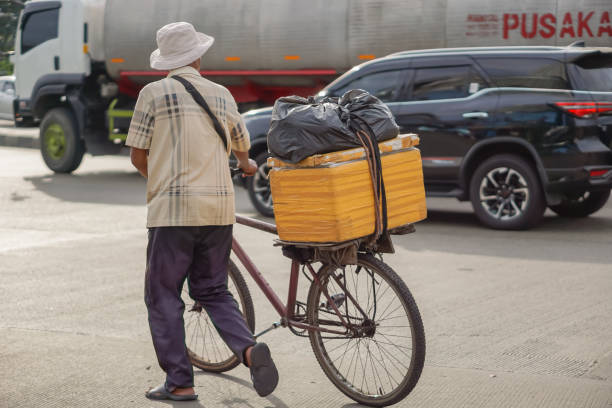 A man wearing a hat walks while guiding his bicycle carrying goods in the back seat in the afternoon on Jalan Prof. Dr. Latumenten, West Jakarta. Jakarta, Indonesia - December 11, 2022.