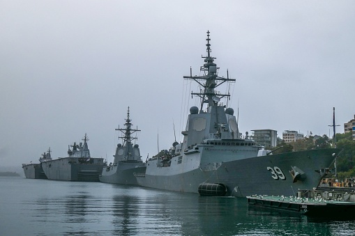 HMAS Hobart (DDG39), a destroyer of the Royal Australian Navy, docked at Garden Island, Sydney Harbour.  Visible behind her are her sister ship, HMAS Sydney (DDG42), and the Amphibious Assault Ships, HMAS Canberra (L02) and HMAS Adelaide (L01).  Seagulls rest on a floating pontoon.   This image was taken on an overcast summer evening.
