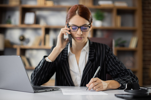 Business, technology, communication and people concept - happy smiling caucasian businesswoman sitting at desk with papers calling on phone at modern office.