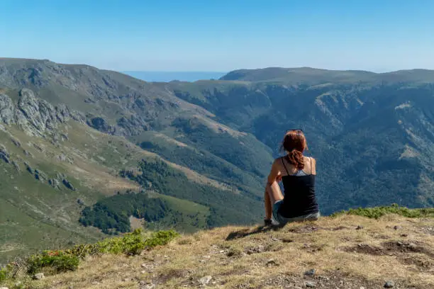 Photo of A woman sitting on top of a mountain in a relaxing mood and looking at a beautiful view of peaks, forests and blue skies