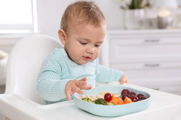lindo bebé comiendo comida en silla alta en casa - baby carrot fotografías e imágenes de stock
