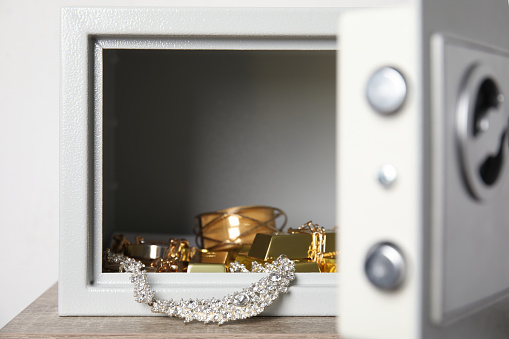 Open steel safe with gold bars and jewelry on wooden table against white background