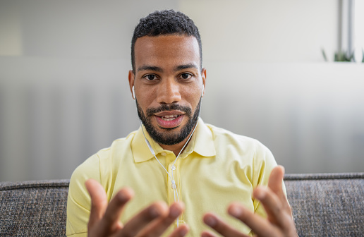 Head shot of african-american man with earphones speaking and looking at camera, chatting online by video call, male vlogger or teacher recording webinar. Man using modern technology for communication. Business and career concept.