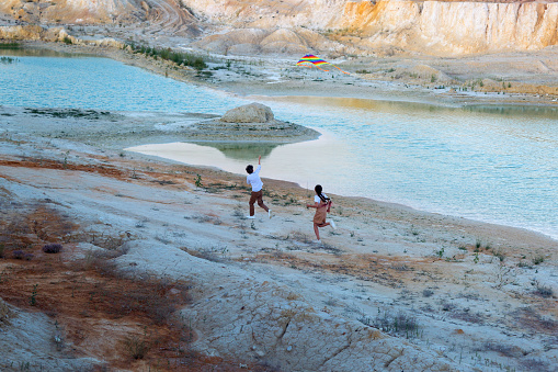 A boy and a girl with a kite are running through a mountainous area along a river.