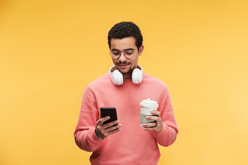 Young multi-ethnic man with white headphones around neck using smartphone and having cup of coffee against yellow background