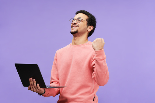 Ecstatic guy in pink casualwear keeping eyes closed while holding laptop and expressing excitement against violet background in isolation