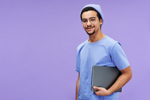 Young smiling businessman or student in blue t-shirt and beanie holding laptop while standing on violet background and looking at camera