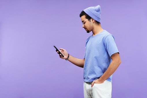Side view of serious guy in blue t-shirt and beanie hat texting in mobile phone or looking through online information on violet background