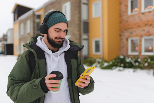 Bearded young man using his phone outdoors while drinking coffee and listening to music.