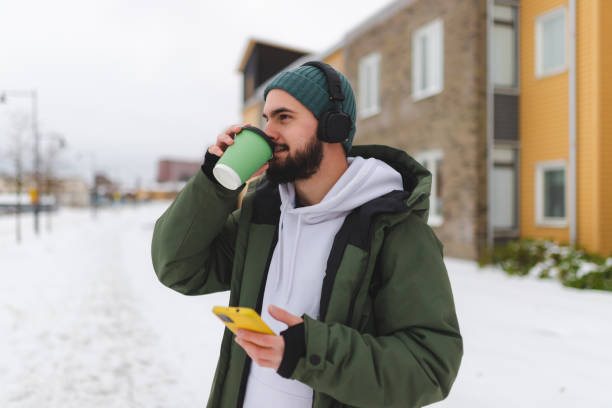 istock Young man with a beard drinking coffee outdoors while using his phone and wearing headphones 1450037272