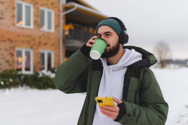 istock Young man with a beard drinking coffee outdoors while using his phone and wearing headphones 1450037317