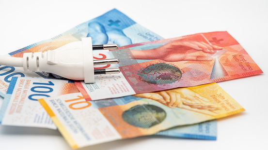 A white J-type power plug with a power cable lies on top of some Swiss banknotes, on a white background. The picture shows banknotes with denominations of 10 francs, 20 francs and 100 francs. Bills of the current 9th series of banknotes. The Swiss Franc is the currency of the Swiss Confederation and the Principality of Liechtenstein. The swiss franc (CHF) is issued by the swiss national bank (SnB).