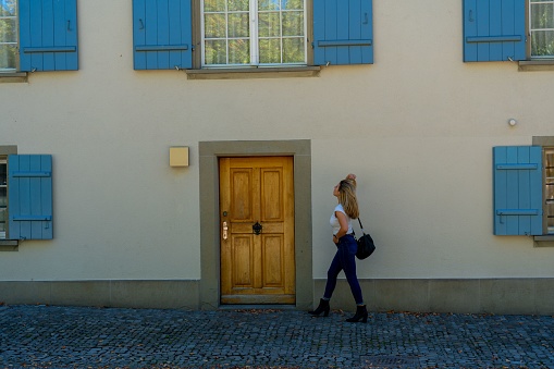 A young blonde woman passing by a building with blue window shutters.
