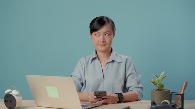 Asian woman using smart phone during working hours sitting in office isolated over blue background.