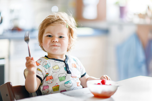 Adorable baby girl eating from spoon fresh healthy raspberries food, child, feeding and development concept. Cute toddler, daughter with spoon sitting in highchair and learning to eat by itself