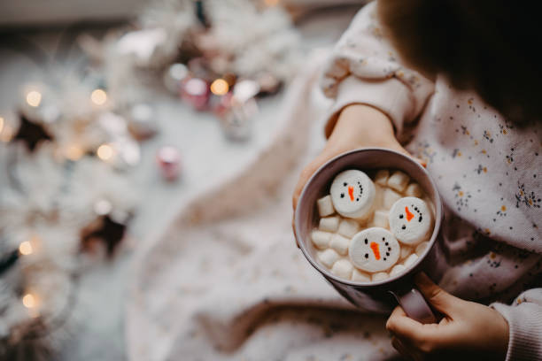 unkenntliches kleines kind, das als schneemann eine tasse mit heißer schokolade mit marshmallows hält. kind sitzt in der nähe eines weihnachtlich geschmückten fensters mit lichtern. - romrodinka stock-fotos und bilder