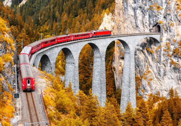 Bernina express glacier train on Landwasser Viaduct in autumn, Switzerland. Scenic view of railroad bridge in orange mountain forest in Swiss Alps. Theme of railway, nature, fall season and travel.