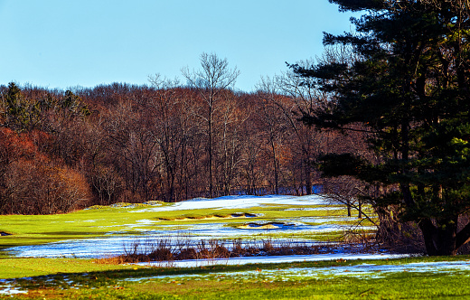 A golf course hole on a sunny day in winter. Snow covers some of the green fairway and all of the putting green, which is protected by sand traps. A forest and lake are in the background.