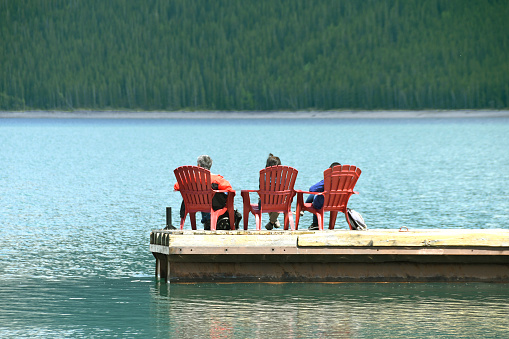 Banff, Alberta, Canada - June 2018: Three people sitting on chairs on a wooden jetty floating on the waters of lake Minnewanka