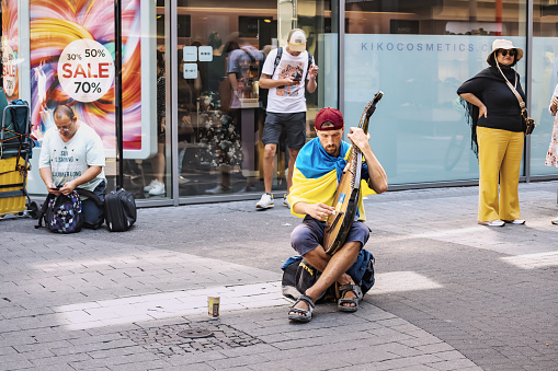 30 July 2022, Cologne, Germany: A refugee from the war in Ukraine, a musician plays bandura national musical instrument on the street of the city, wrapped in the Ukrainian flag