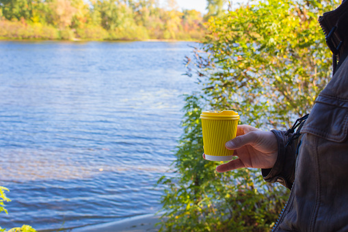 Man holding yellow cup on river background. Coffee cup in nand on autumn landscape background. Travel concept. Vacations at nature. Coffee to go concept. Picnic at the riverbank. Active morning.
