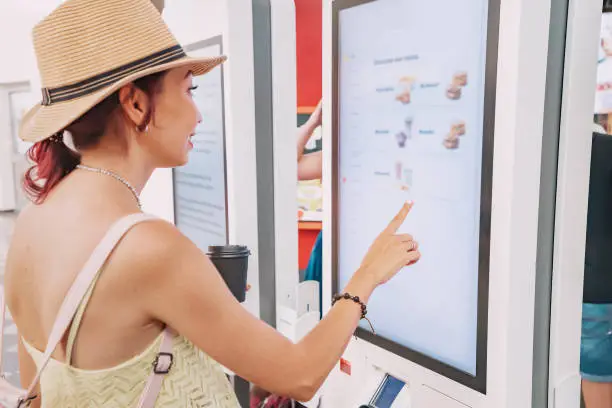Photo of A female customer uses a touchscreen terminal or self-service kiosk to order at a fast food restaurant. Automated machine and electronic payment
