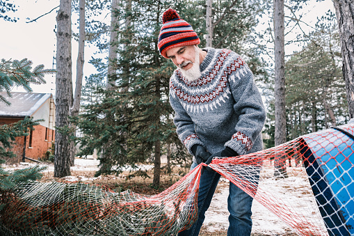 Christmas tree hunt in nature, Mature man with beard packing  freshly cut Christmas tree with wire wrapper.   He is wearing knit sweater and hat with jeans. Outdoor of the farm and wooded area in winter.