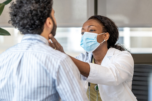 Black female doctor with surgical mask checking a young middle-eastern man's lymph nodes in her office.
