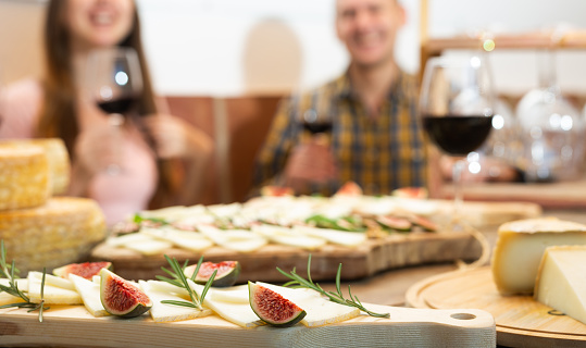 One man and two women enjoying degustation of cheese while holding glasses with red wine on rustic wooden table in restaurant