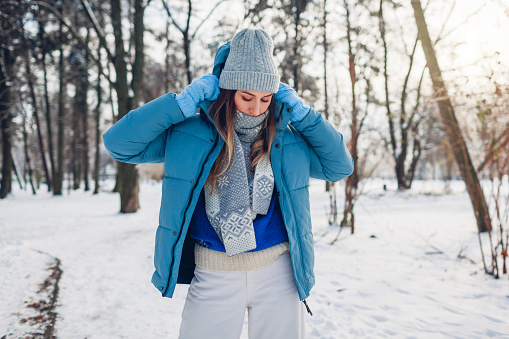 Portrait of young woman putting on blue coat walking in snowy winter park. Warm clothes for cold weather. Sweater season