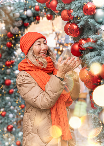 Asian woman in orange scarf and hat walking in Christmas market decorated with holiday lights. Feeling happy in  city Christmas decorated.