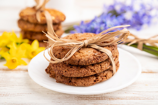 Oatmeal cookies with spring snowdrop flowers bluebells, narcissus and cup of coffee on white wooden background. side view, close up, defocused, still life. Breakfast, morning, spring concept.