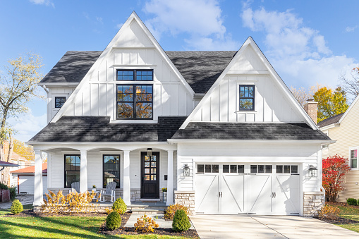 Oak Park, IL, USA - November 4, 2020: A new, white modern farmhouse with a dark shingled roof and black window frames. The bottom of the house has a light rock siding and covered front porch.