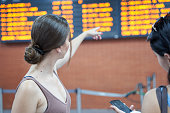 Back view of couple in airport, looking and pointing at travel board with flights information and checking their smartphone