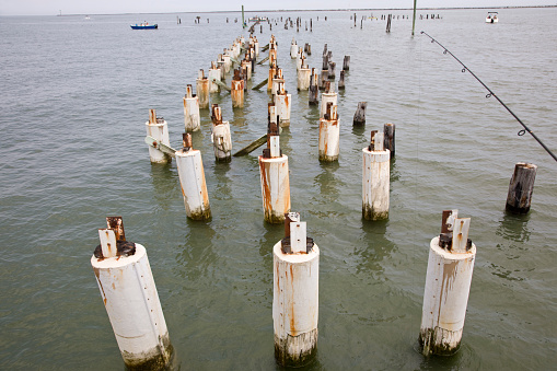 Deteriorated old pier pilings with small boats in the background at the shoreline of Delaware Bay USA.