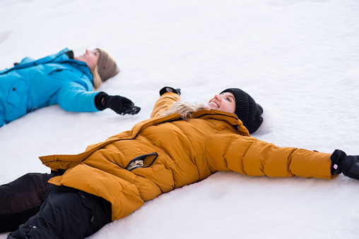 Two beautiful young women wearing warm winter coats and hats are laying in the snow together with their arms spread making snow angels playfully in the freshly fallen snow.
